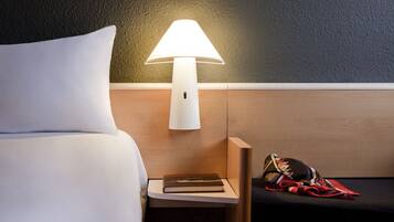 Ein ordentlich bezogenes Bett mit einem weißen Kopfkissen, ein Nachttisch mit einer Lampe, einem Buch und einem roten und schwarzen Gegenstand mit Muster.