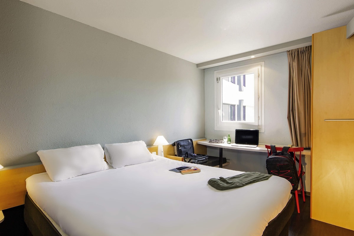 Quarto de hotel com cama, escrivaninha com um laptop, cadeira e mala.