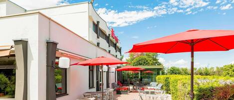 An outdoor seating area with tables and chairs, red umbrellas, and a building with a white facade.