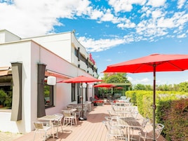 An outdoor seating area with tables and chairs, red umbrellas, and a building with a white facade.