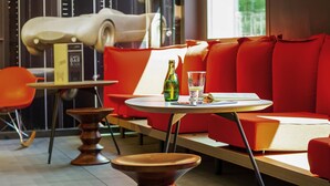 A modern interior with a red sofa, a round table with a bottle and glass, and a book. - ibis Mulhouse Ile Napoléon (Sausheim)