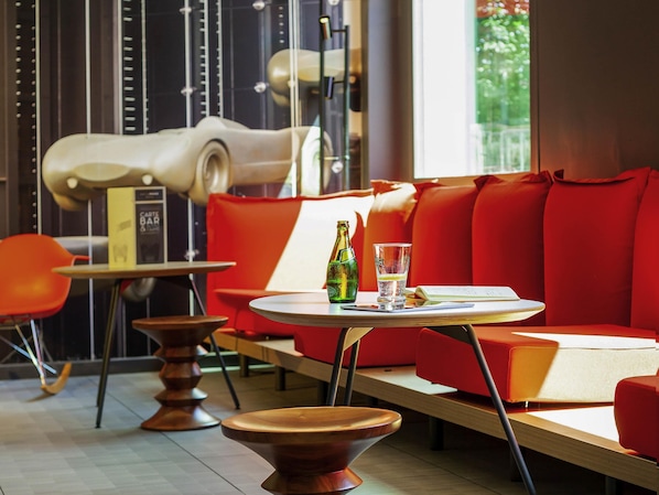 A modern interior with a red sofa, a round table with a bottle and glass, and a book. - ibis Mulhouse Ile Napoléon (Sausheim)