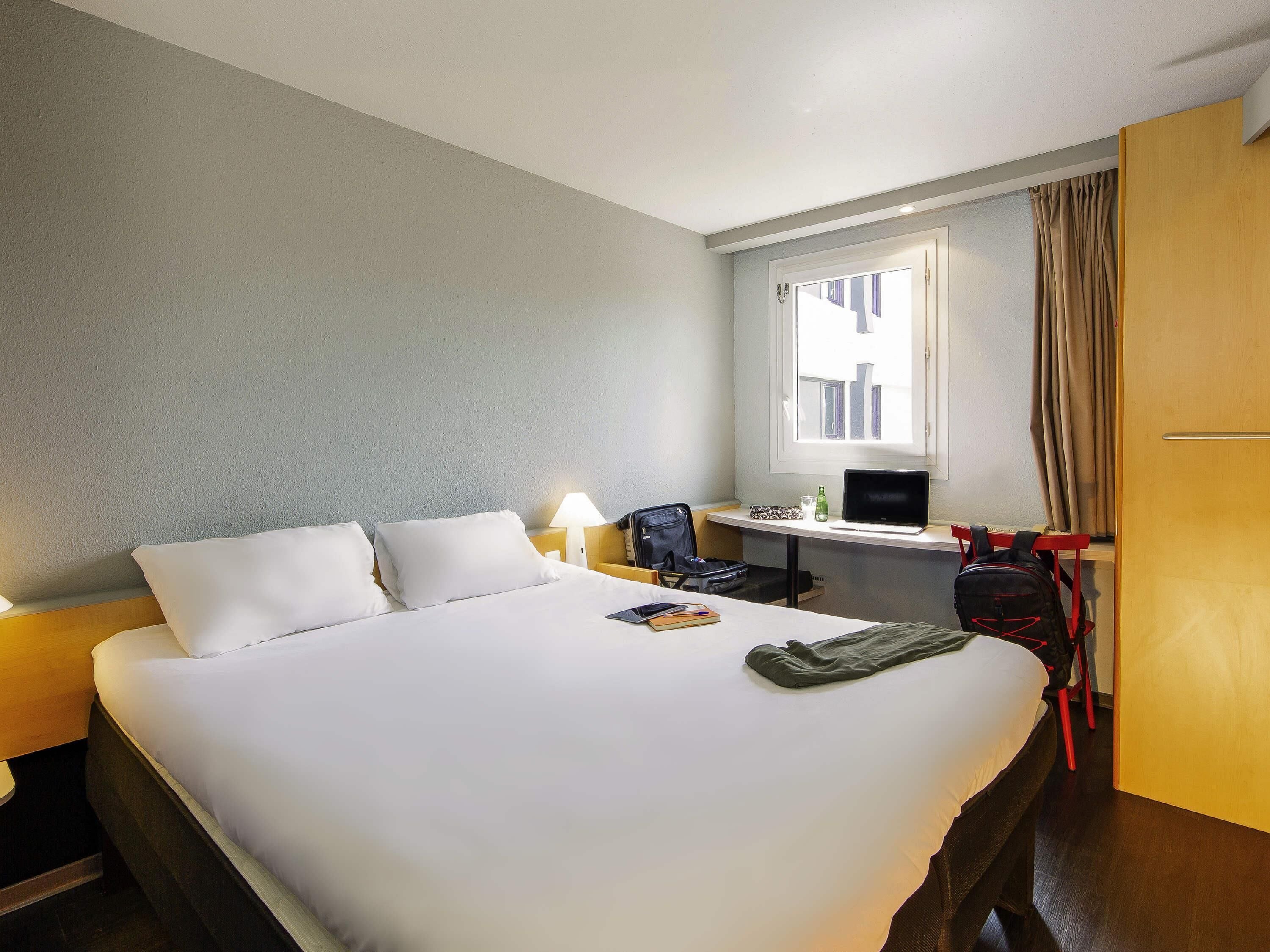 A hotel room with a bed, a desk, a chair, a laptop, a backpack, and a window with curtains.