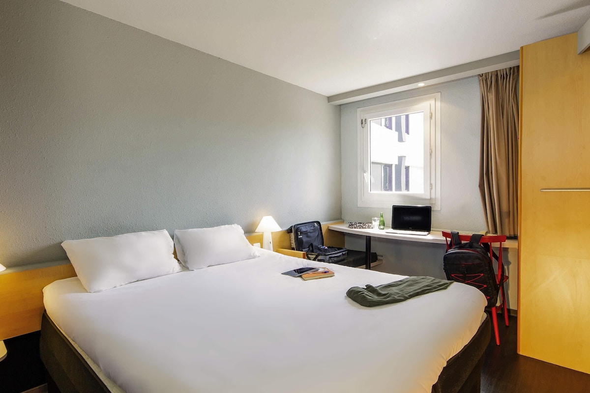 A hotel room with a bed, a desk, a chair, a laptop, a backpack, and a window with curtains.