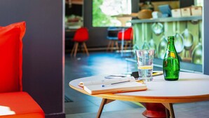 A table with a notebook, a glass, and a green bottle in a modern interior with red chairs and a bar area. - ibis Mulhouse Ile Napoléon (Sausheim)