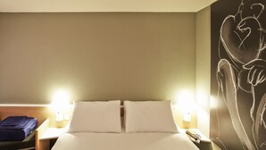 A hotel room with a bed, two bedside lamps, a nightstand, a blue suitcase, and a wall with a chalkboard drawing. - ibis Mulhouse Ile Napoléon (Sausheim)