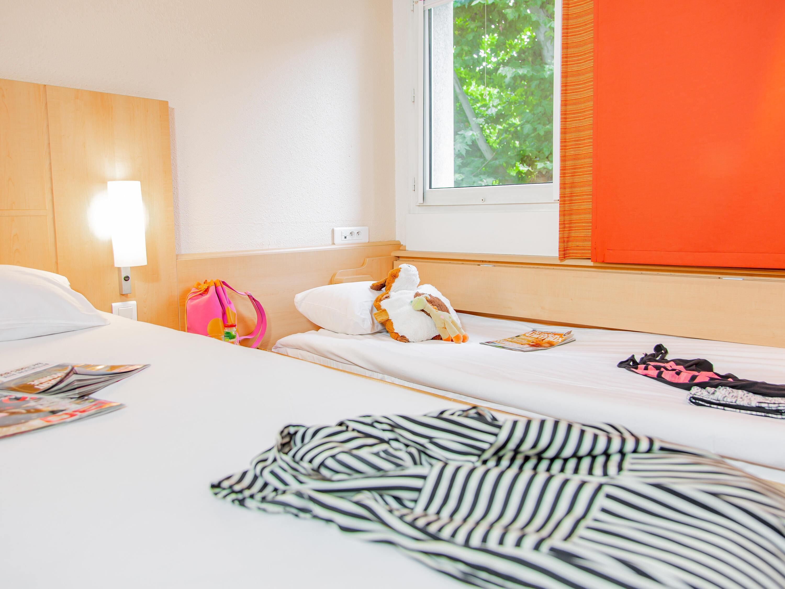 A hotel room with a bed, a window with blinds, a lamp, a stuffed toy, a pink bag, and a magazine.