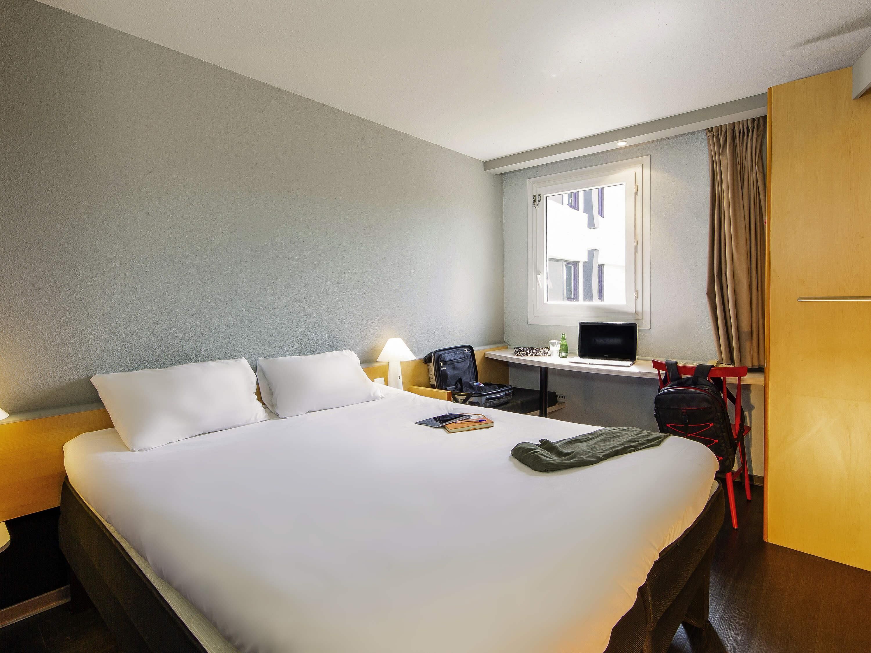 A hotel room with a bed, a desk, a chair, a laptop, a backpack, and a window with curtains.