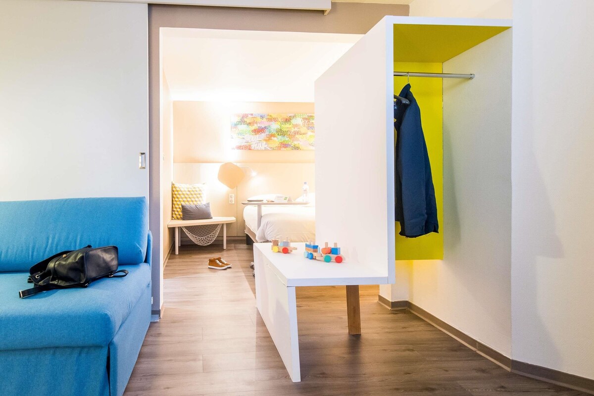 A hotel room with a blue sofa, a small table with toys, a hanging coat rack, and a bed in the background.