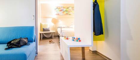 Habitación de hotel con un sofá azul, una mesita con juguetes, un perchero y una cama al fondo.