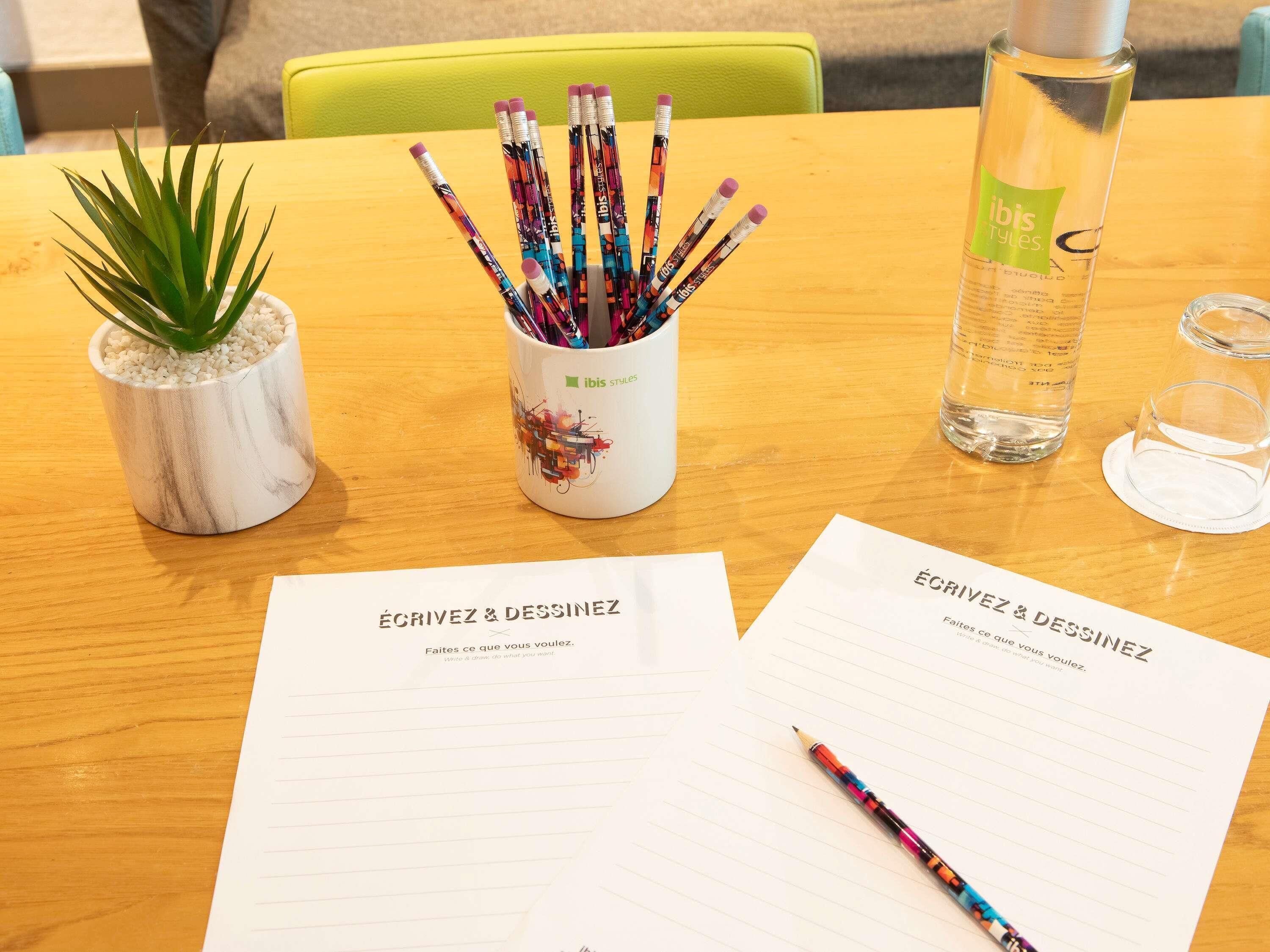 A wooden table with a potted plant, a mug with writing utensils, a water bottle, and two sheets of paper with the text 'ÉCRIVEZ & DESSINEZ'.