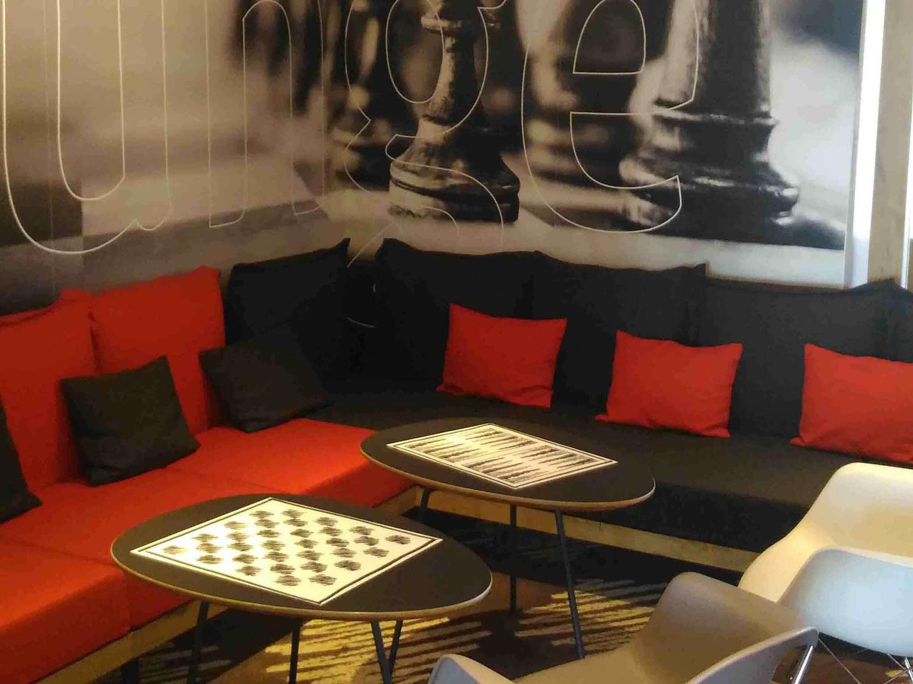 Un área de descanso moderna con un sofá rojo, cojines negros y rojos, y dos mesitas con folletos informativos.