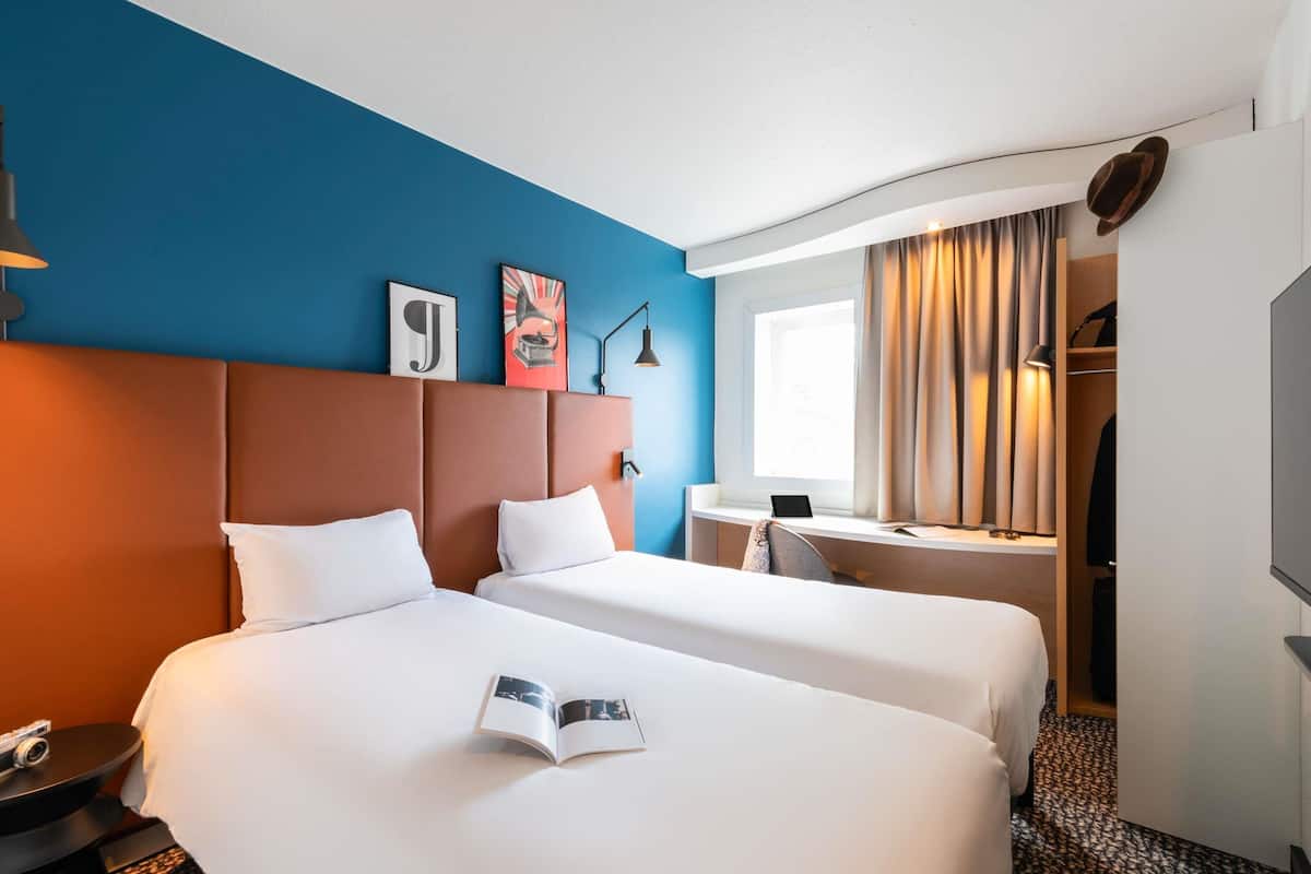 A hotel room with two beds, a desk, a television, and a window with curtains.