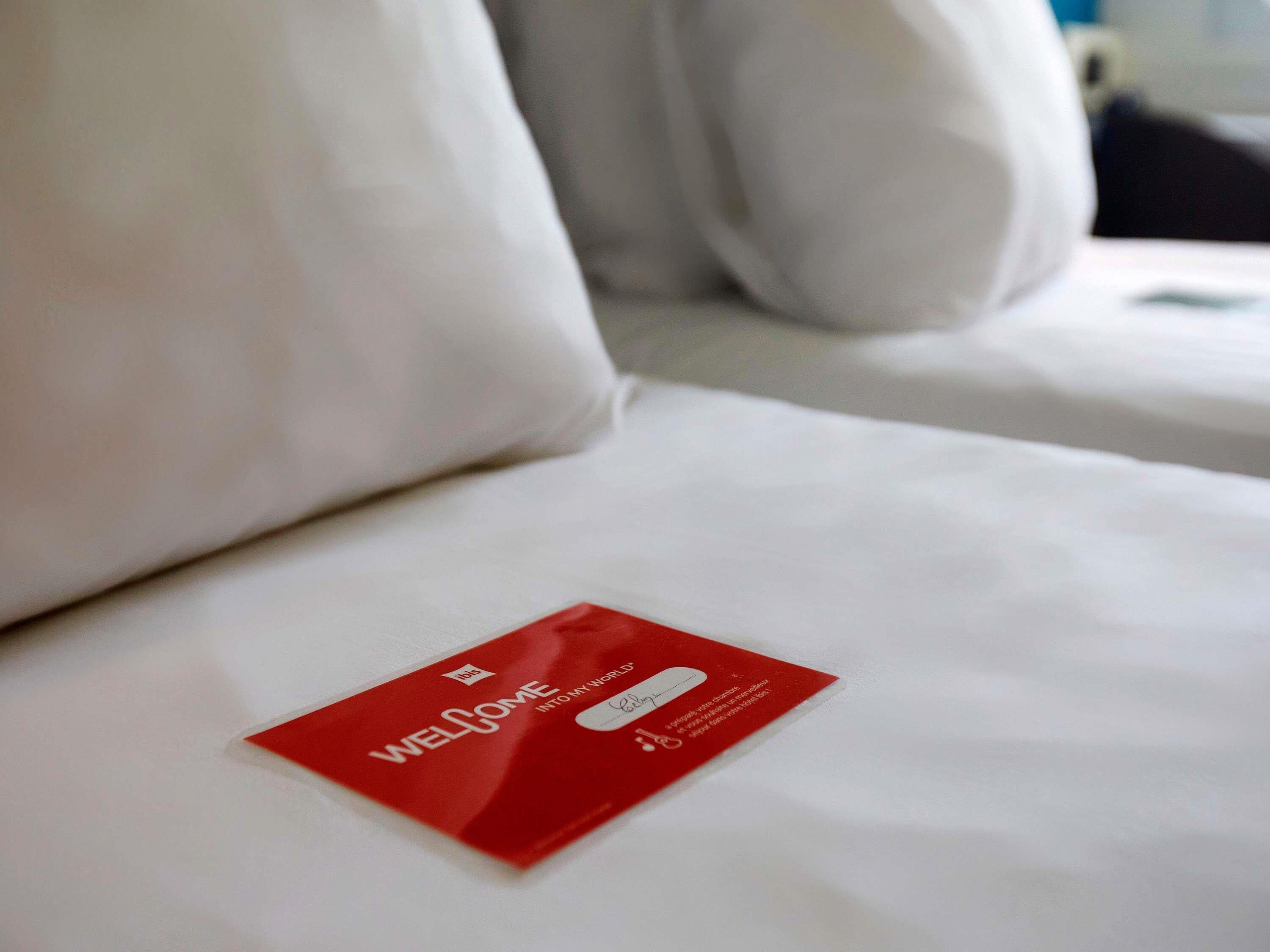 Habitación de hotel con una cama bien hecha, una tarjeta de bienvenida roja sobre la cama y ropa de cama blanca.