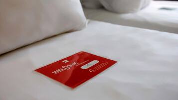 Habitación de hotel con una cama bien hecha, una tarjeta de bienvenida roja sobre la cama y ropa de cama blanca.