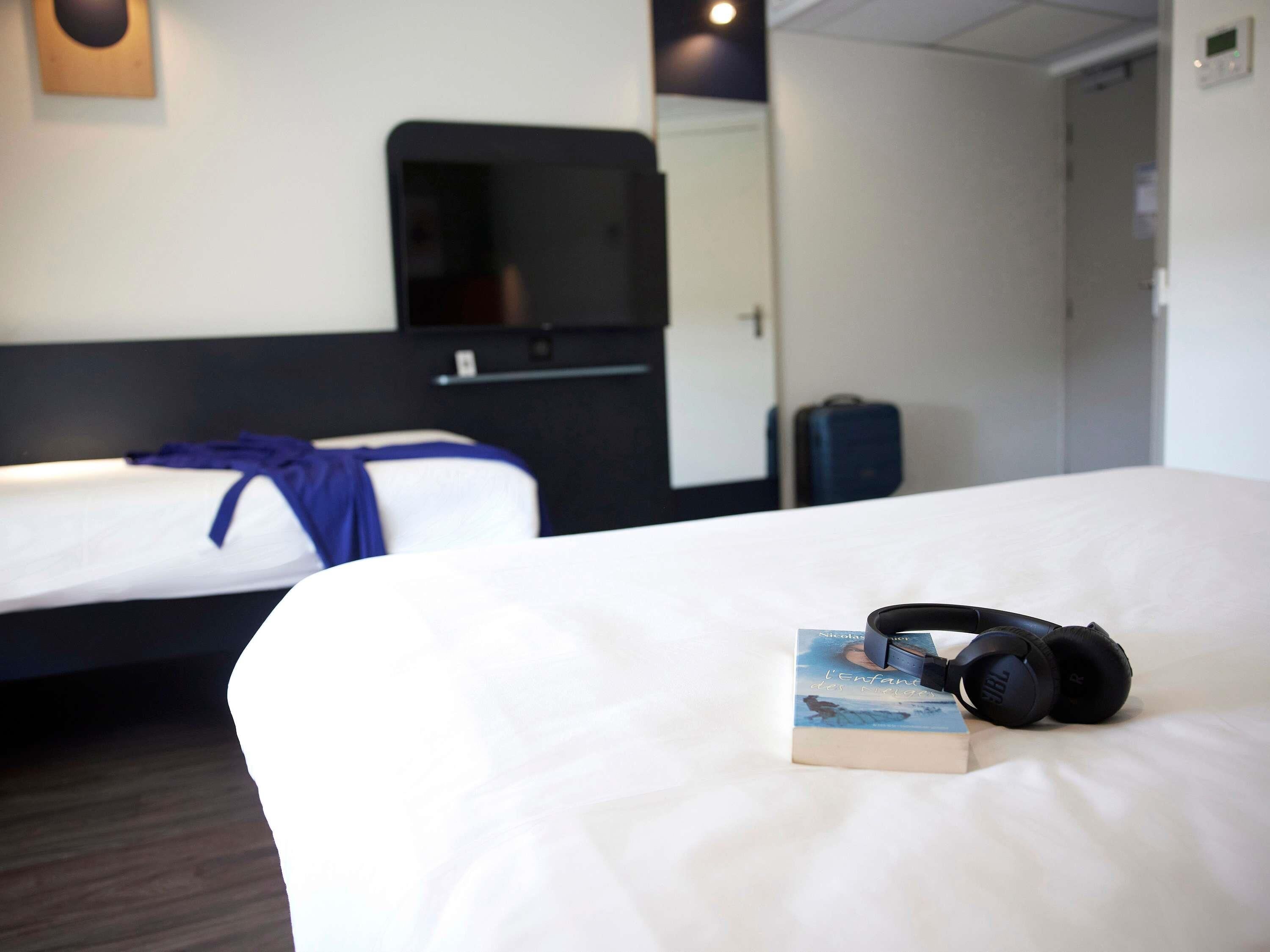 Una habitación de hotel con una cama, un libro, audífonos, una maleta y un televisor.