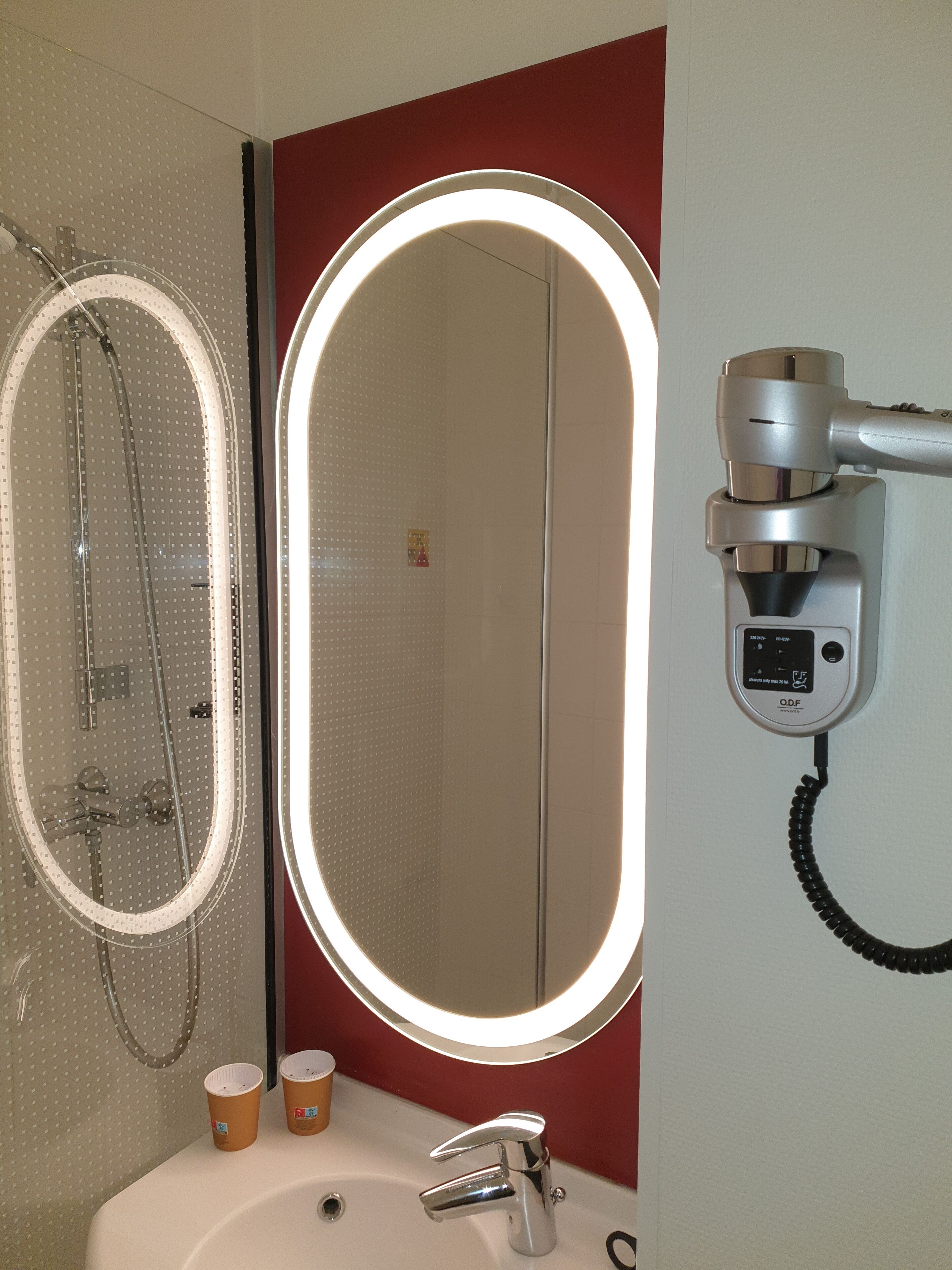 Une salle de bain avec un miroir rond, un sèche-cheveux et deux gobelets en papier posés sur le lavabo.