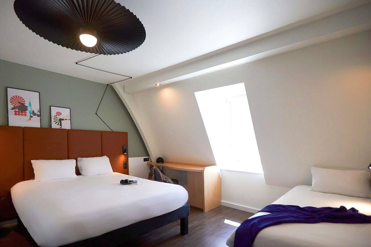 A modern hotel room with a large bed, a smaller bed, a desk, and a window.
