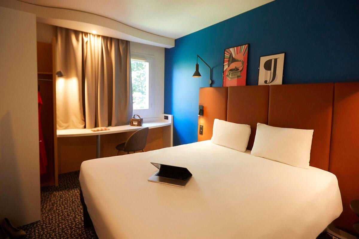 A hotel room with a bed, a desk, a chair, a laptop, and a window with curtains.