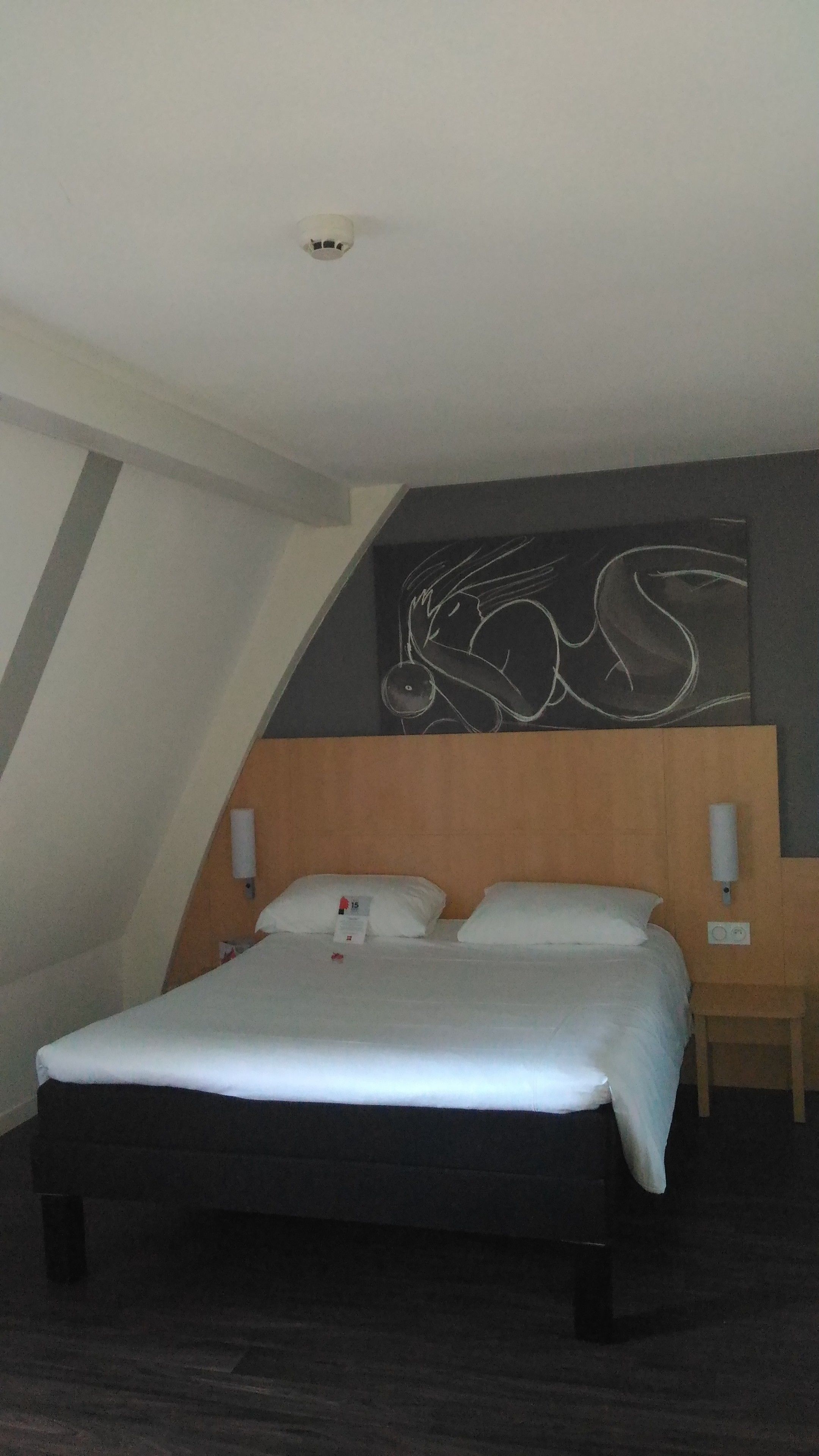 A hotel room with a large bed, a wooden headboard, a wall-mounted artwork, and a small bedside table.