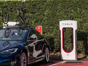 Ein Tesla Elektroauto geparkt neben einer Tesla Ladestation, im Hintergrund ein Hecken.