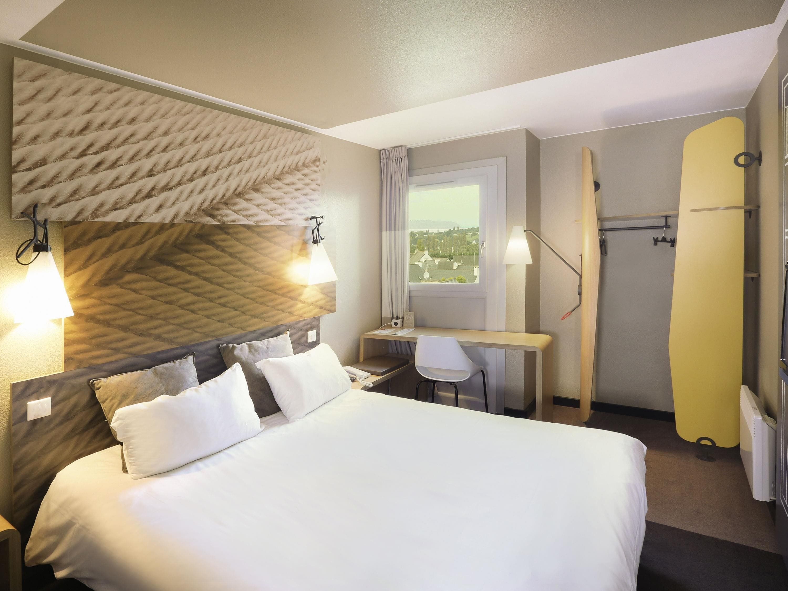 A hotel room with a bed, desk, chair, and a view of the outside.