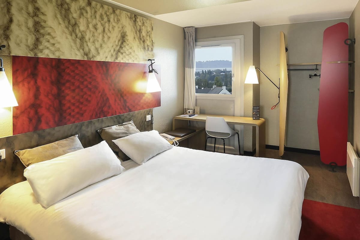 A hotel room with a bed, desk, chair, and a red snowboard leaning against the wall.