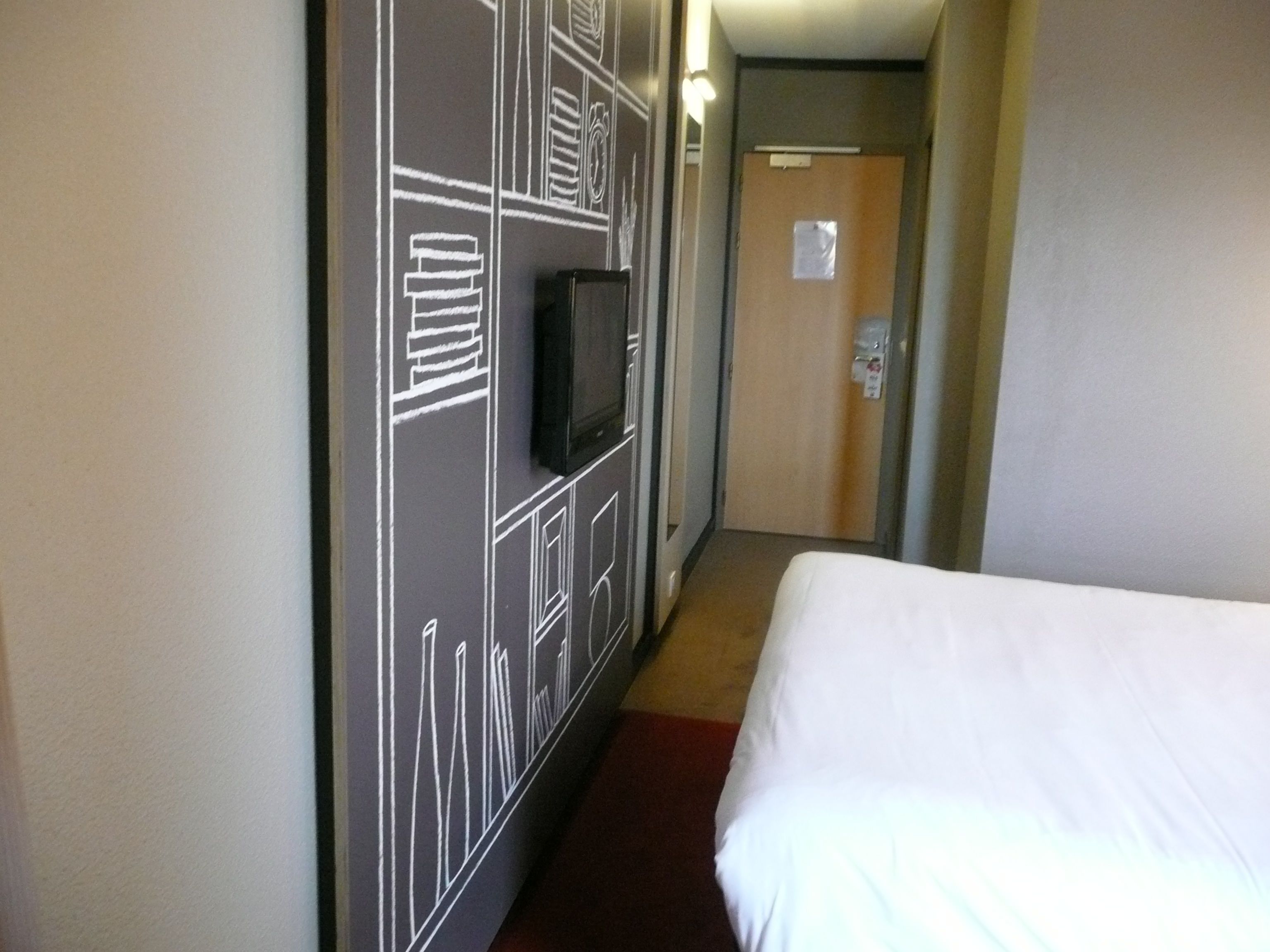 Ein Hotelzimmer mit einem Bett, einem an der Wand befestigten Fernseher und einer Tür am Ende des Ganges.