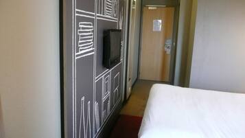 Ein Hotelzimmer mit einem Bett, einem an der Wand befestigten Fernseher und einer Tür am Ende des Ganges.
