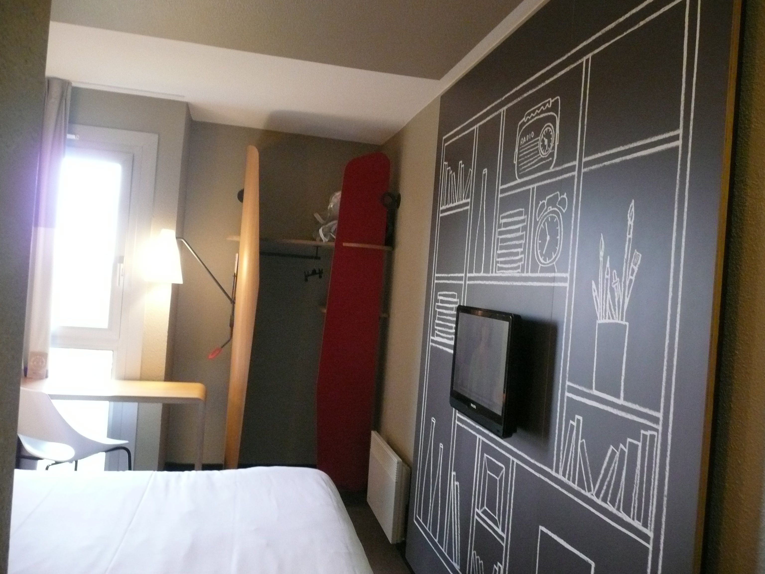 A hotel room with a bed, a desk, a chair, a TV mounted on the wall, and a red suitcase.