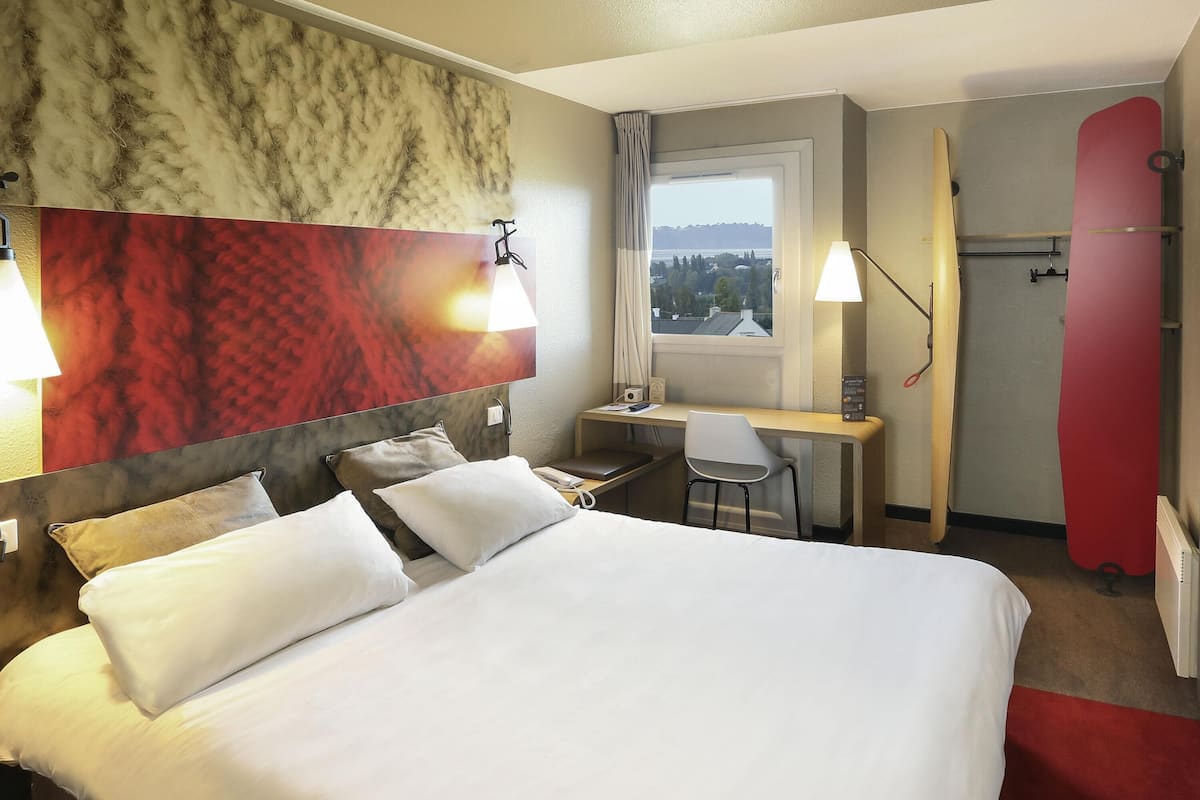 A hotel room with a bed, desk, chair, and a surfboard.