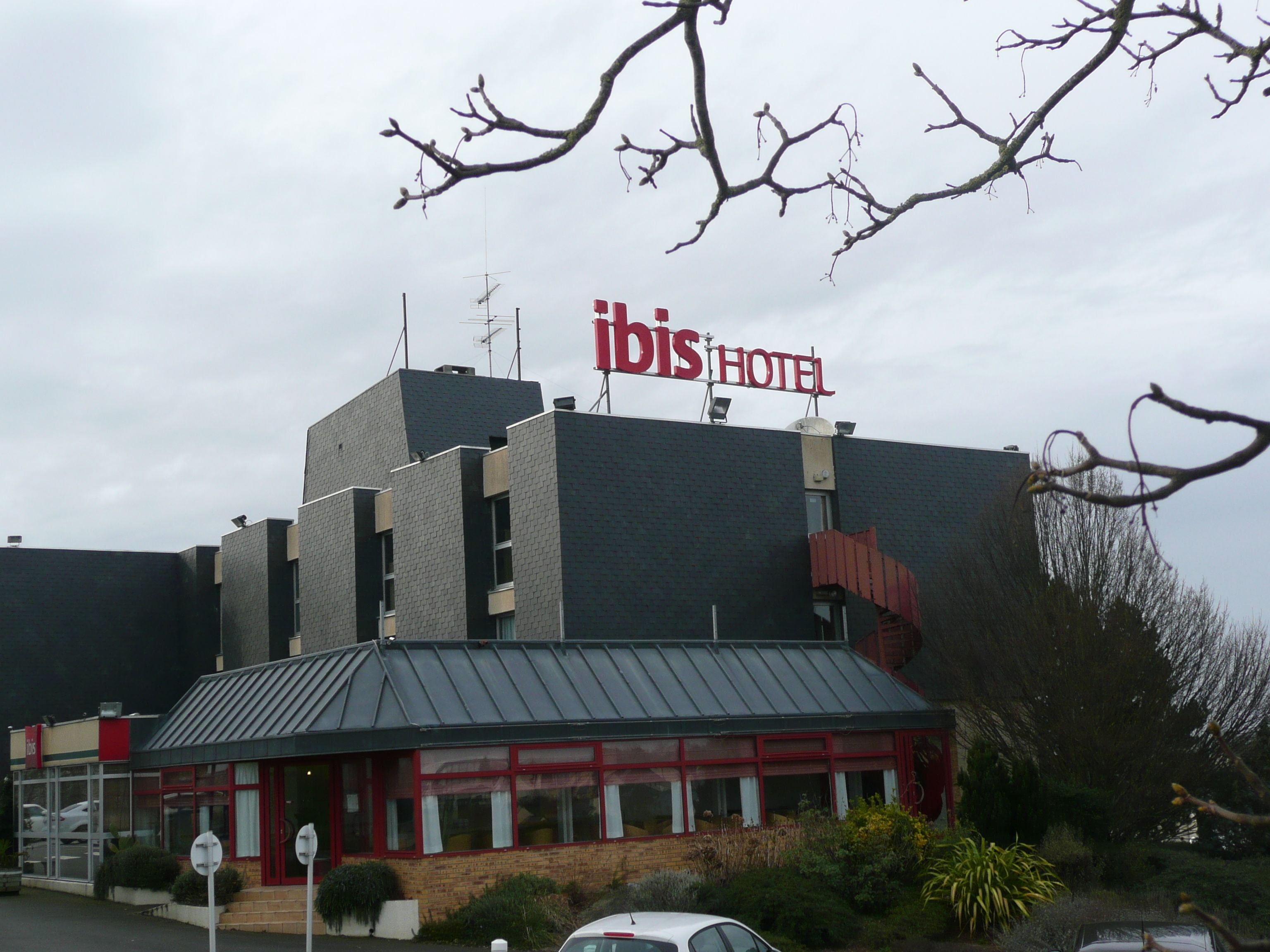 A hotel with a modern architectural design, featuring a prominent sign for 'ibis HOTEL' and a parking area with cars.