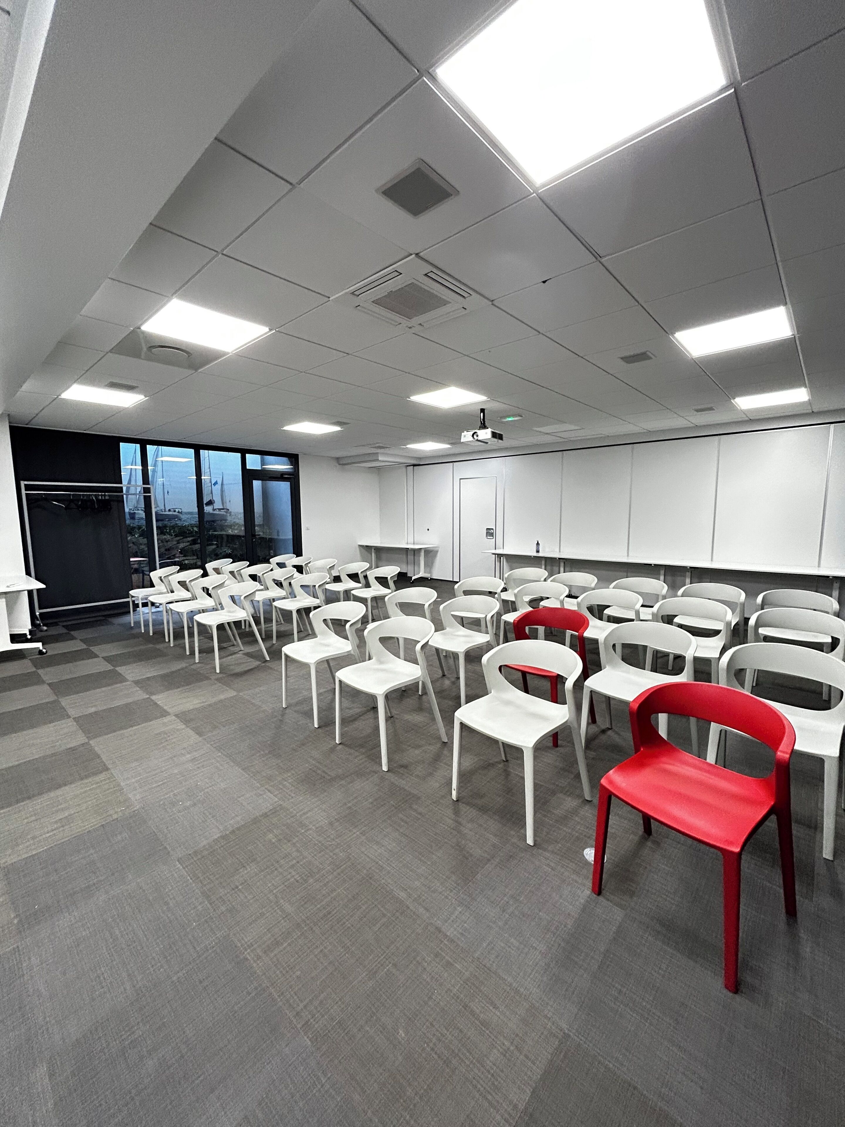 A conference room with rows of white chairs, a projector, and a large window with a view of sailboats.