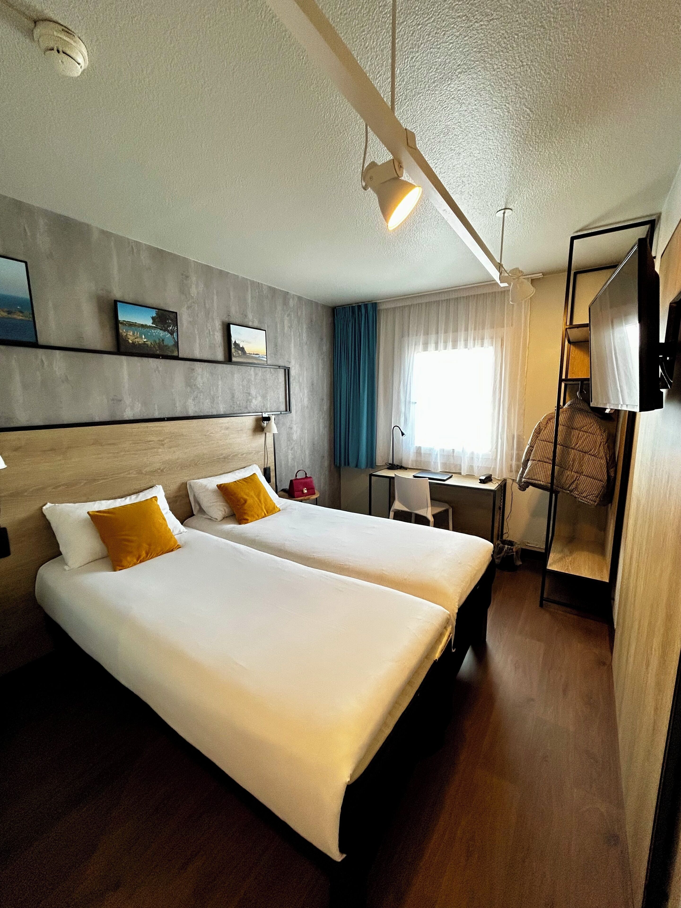 A hotel room with two beds, a desk, a chair, a TV, and a window with curtains.