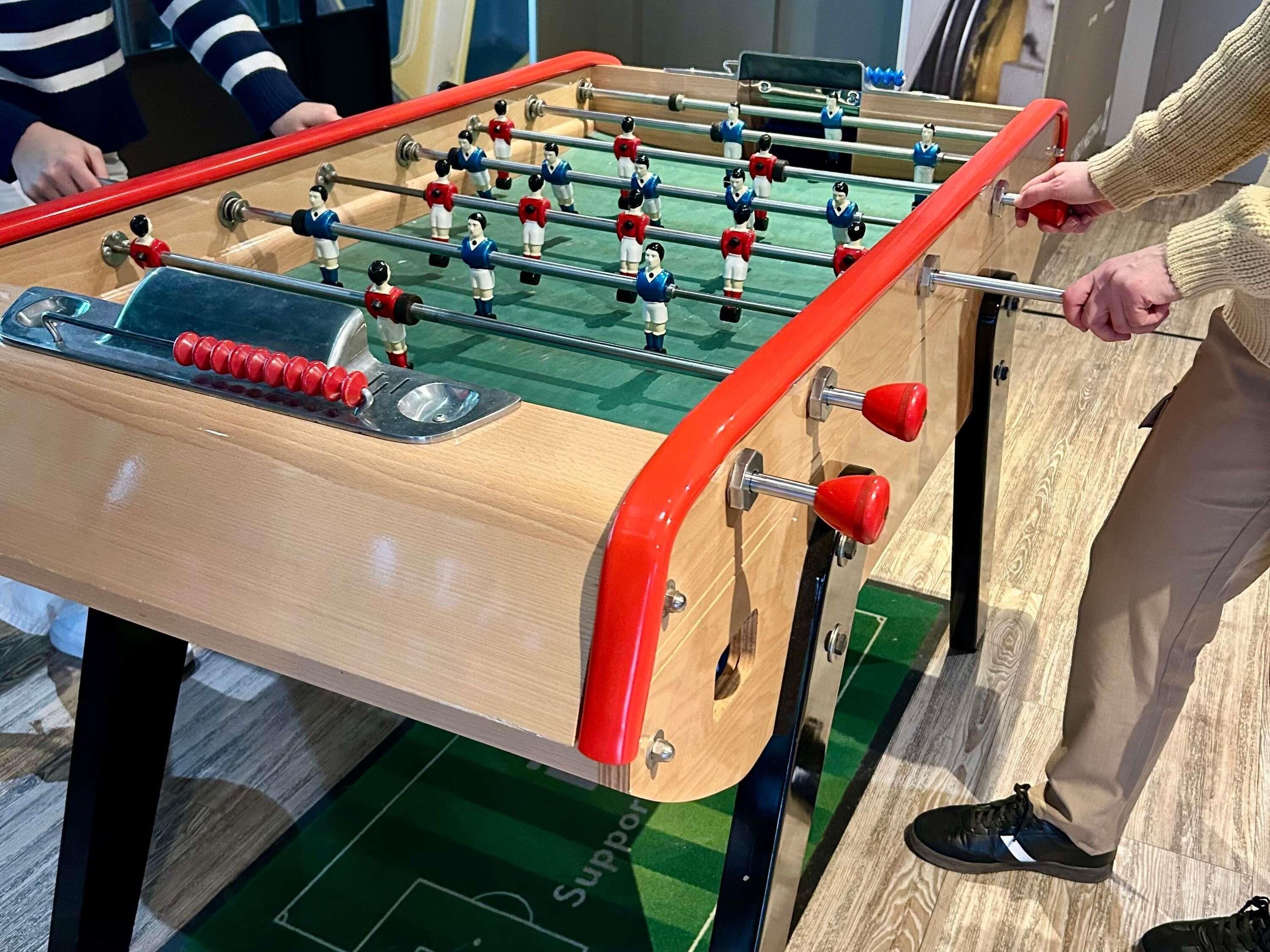 Ein Holztischfußball mit roten Griffen und Metallstangen, platziert auf einem grünen Boden mit Fußballfeld-Muster.