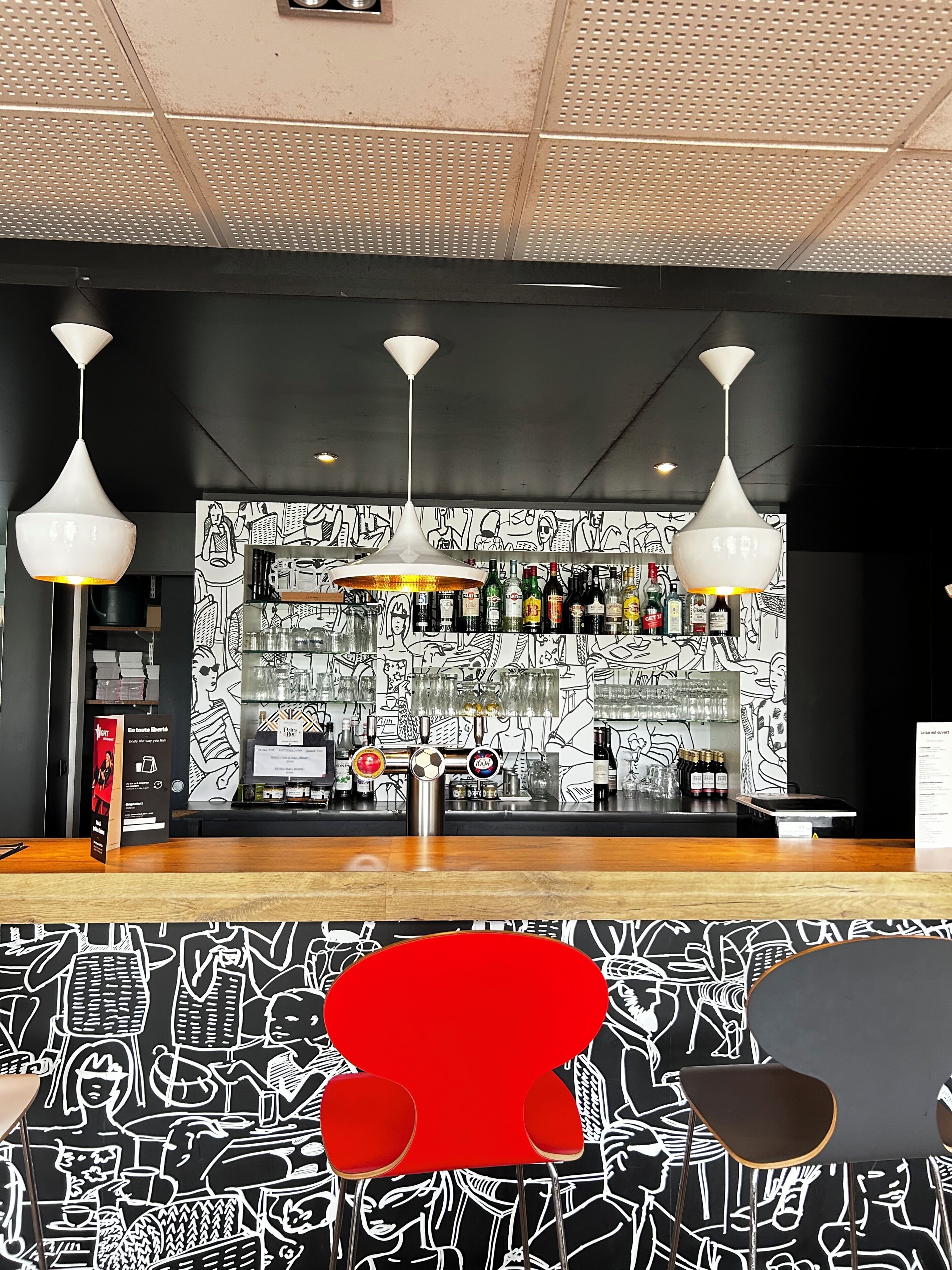 A bar area with a black and white mural, three pendant lights, and a variety of bottles and glasses on shelves.