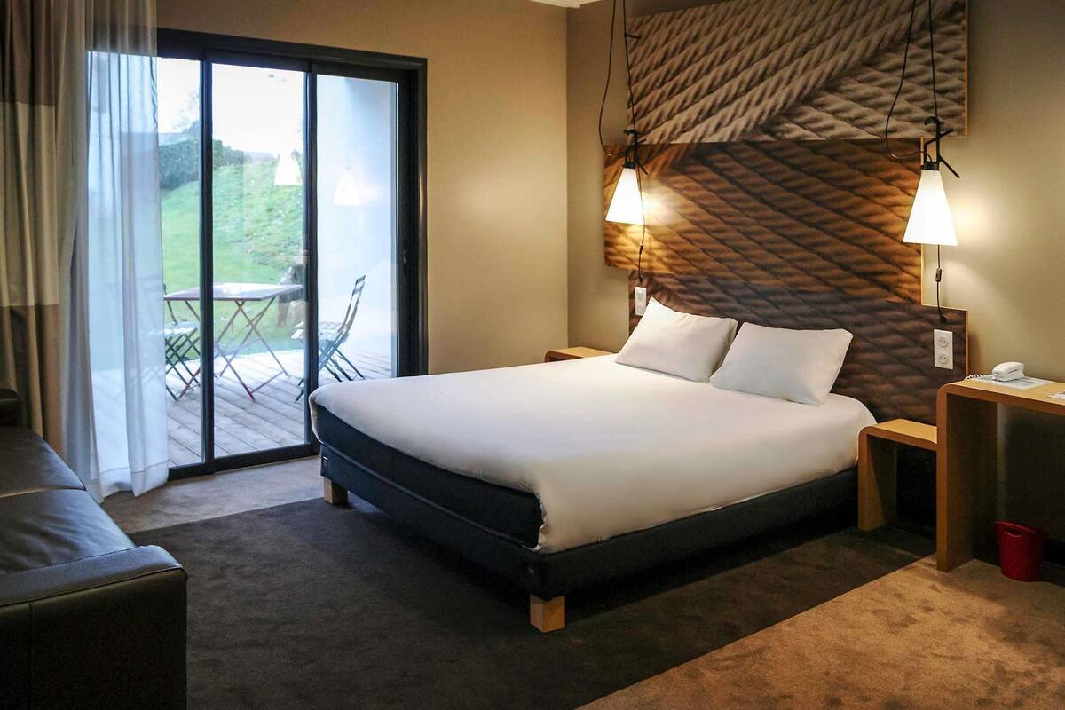 A hotel room with a large bed, a wooden headboard, a desk with a phone, and a view of a deck through a sliding glass door.