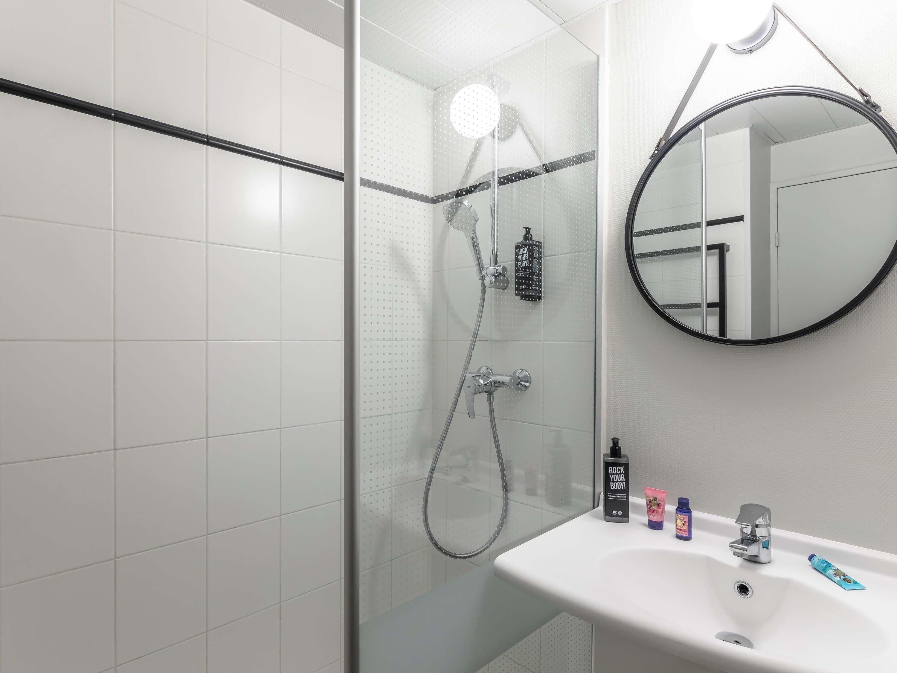 Ein modernes Badezimmer mit einer Duschkabine aus Glas, einem runden Spiegel und einem weißen Waschbecken mit Pflegeprodukten.
