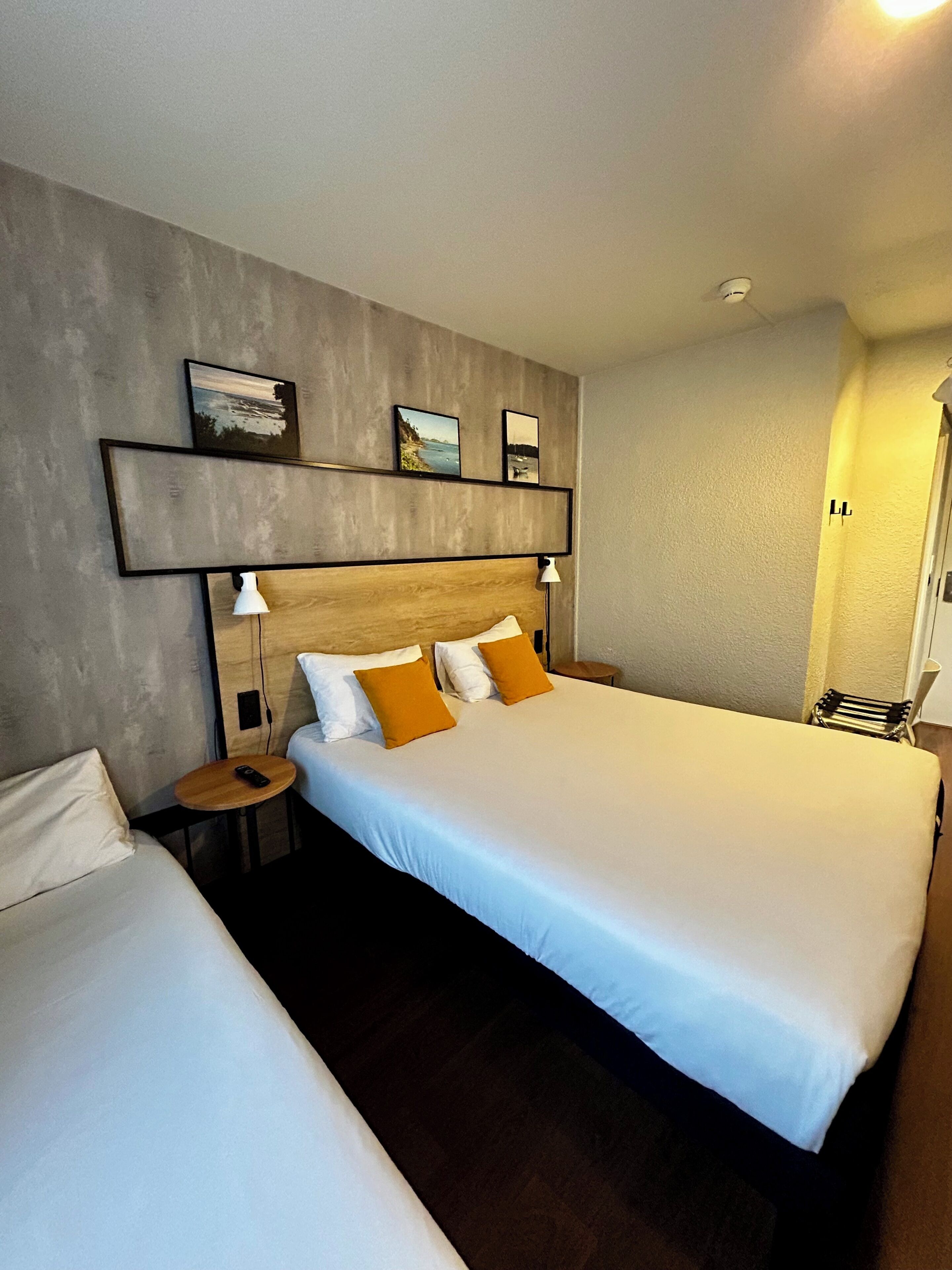 Una habitación de hotel con una cama grande, una cama más pequeña, una mesita de noche, una lámpara y un estante en la pared con fotografías.