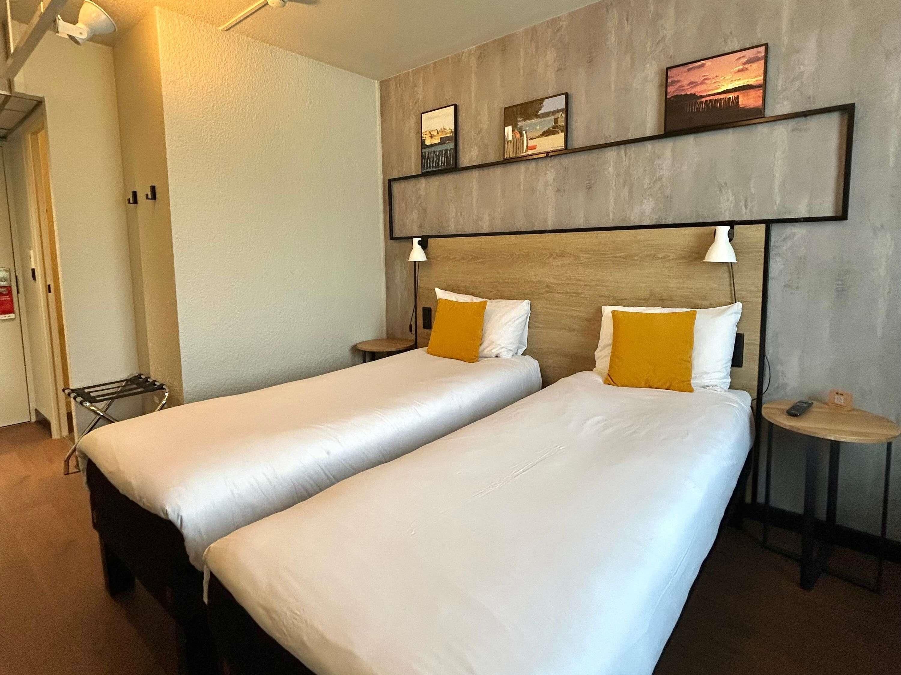 Quarto de hotel com duas camas, cabeceira com obra de arte, luminárias de cabeceira e uma mesinha com telefone.