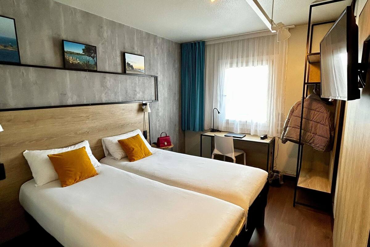 A hotel room with two beds, a desk, a chair, a TV, and a window with curtains.