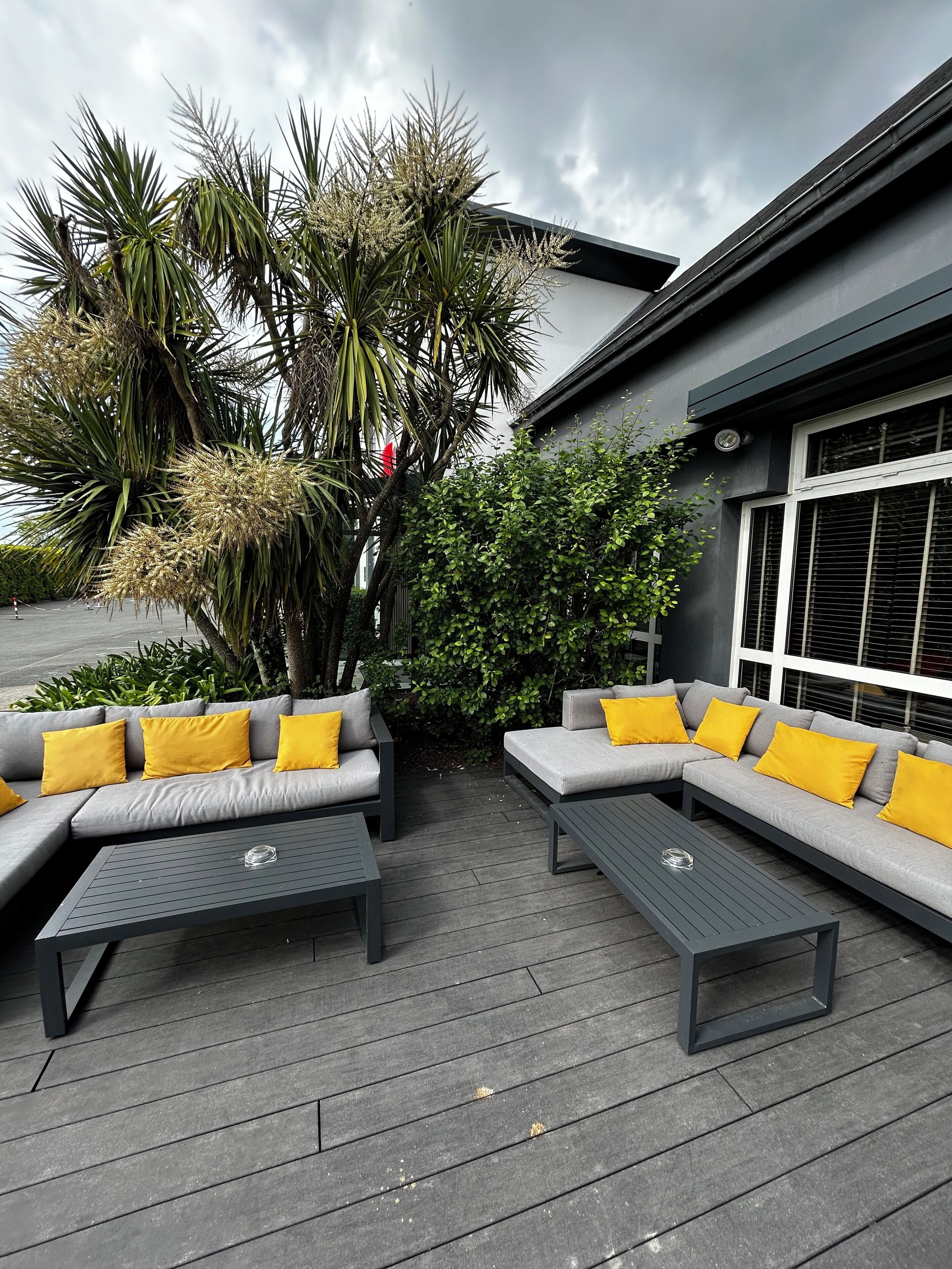 An outdoor seating area with grey sofas and yellow cushions, surrounded by greenery and a modern building.