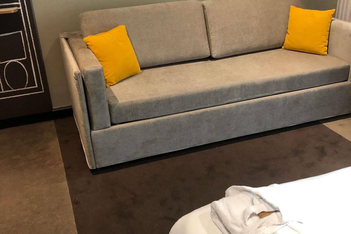 Un sofá gris con dos almohadones amarillos, una cama blanca con una manta blanca y un suelo cubierto de alfombra marrón.