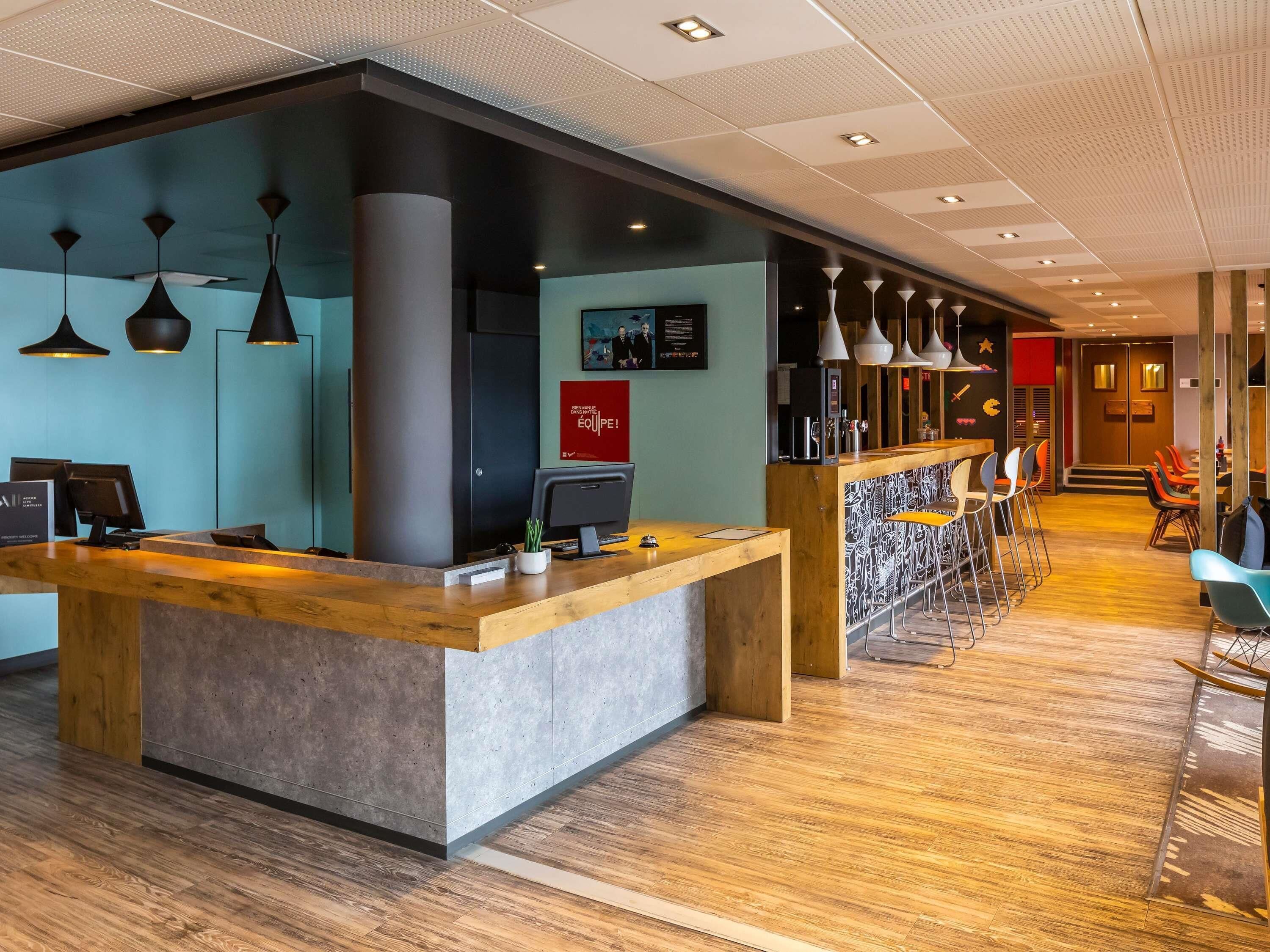 Een modern hotel ontvangstgebied met een houten balie, een digitaal scherm en een zithoek met barstoelen.