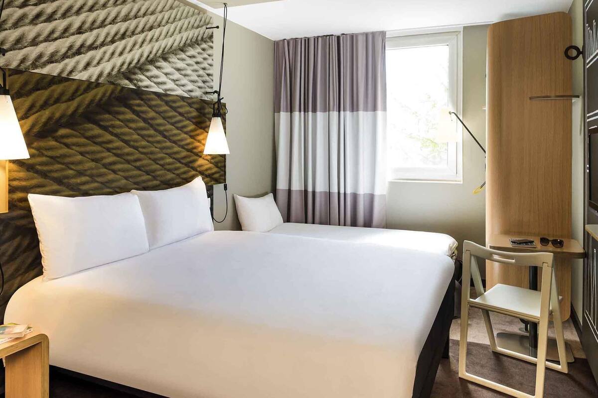 Quarto de hotel com uma cama grande, uma mesinha de cabeceira, uma escrivaninha, uma cadeira, cabeceira com design texturizado, uma janela com cortinas e uma luminária de parede.