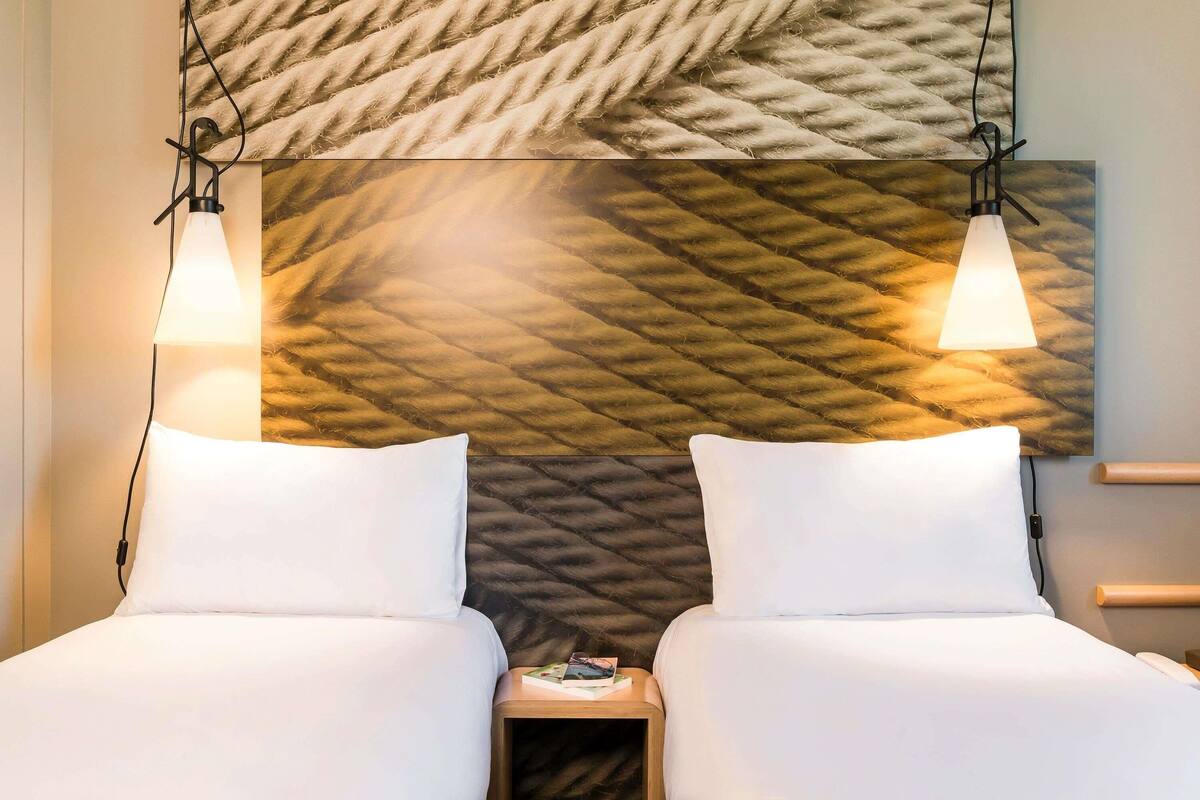 Quarto de hotel com duas camas, parede texturizada e duas luminárias pendentes.