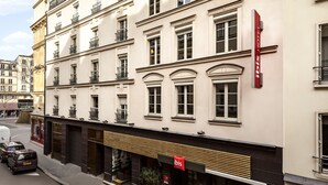Un hôtel à la façade classique, avec une enseigne « ibis » bien visible, de grandes fenêtres et une entrée moderne.