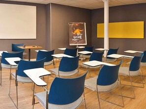 Een klaslokaal met blauwe stoelen, witte tafels, een projectorscherm en een poster aan de muur.