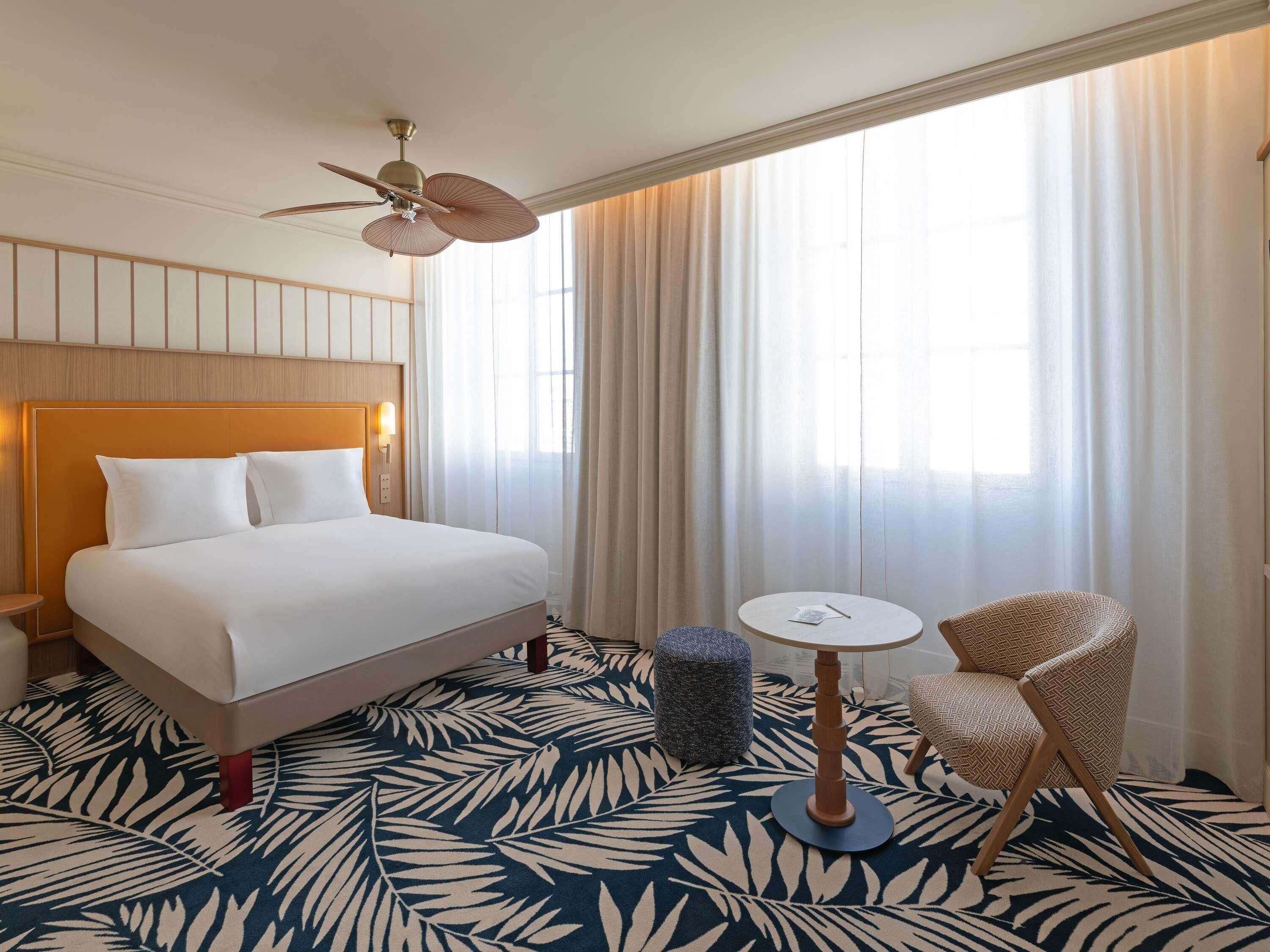 Habitación de hotel con cama, ventilador de techo, una mesita, una silla y una ventana con cortinas.