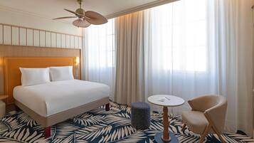 Habitación de hotel con cama, ventilador de techo, una mesita, una silla y una ventana con cortinas.